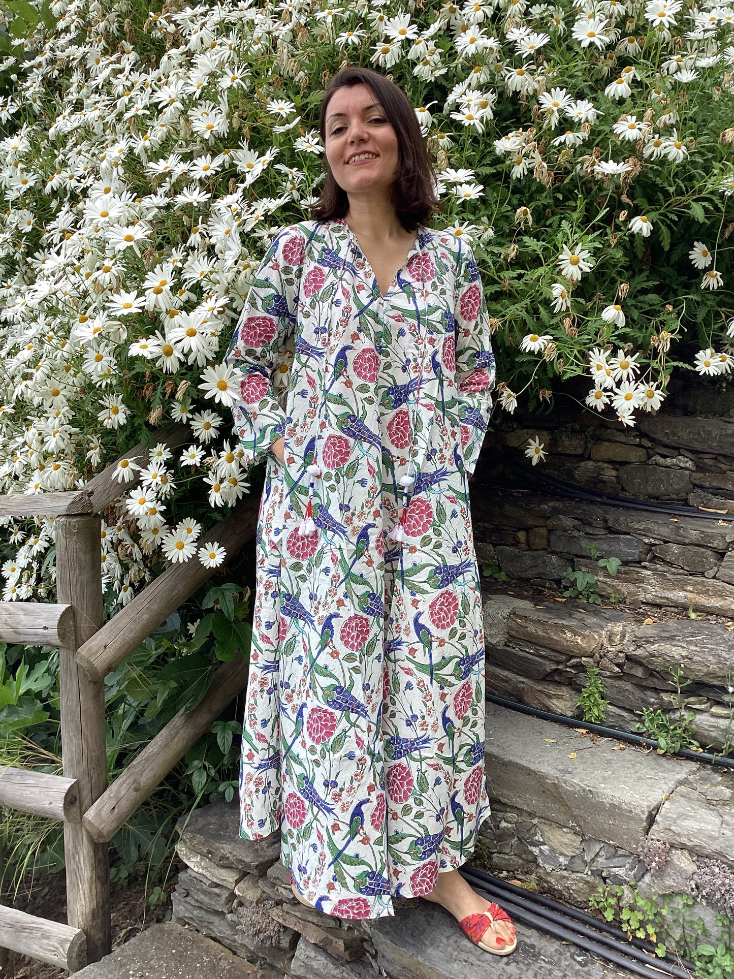 Woman in a floral dress standing in a garden with white flowers and stone wall.